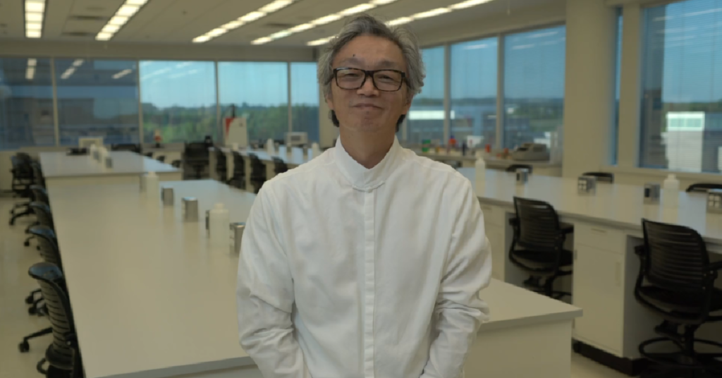 Dr. Jian Han stands facing the camera in a polished white shirt, slightly smiled. Slightly blurred for the background is the view of an empty lab room at Hudson Alpha Institute for Biotechnology